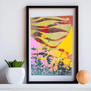 May include: Abstract art print in a black frame. The artwork features a colorful landscape with pink, yellow, green, and red hues. Silhouetted plants are in the foreground. A white pot with a green plant and a wooden sphere are on the white surface.