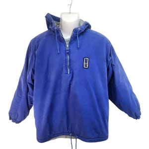 May include: A blue anorak jacket with a hood and a half-zip. It has a small logo patch on the chest and a drawstring hem. The jacket is displayed on a mannequin, suitable for outdoor wear.