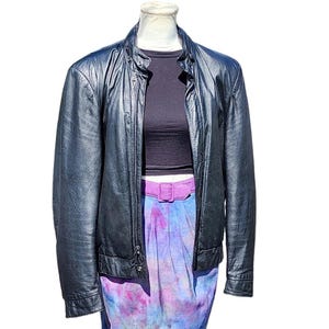May include: A black leather motorcycle jacket with a zipper closure and a stand-up collar. The jacket is worn over a black crop top and a tie-dye skirt with a purple belt.