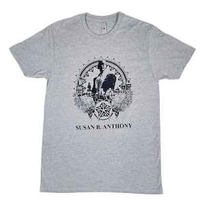 May include: A heather grey t-shirt with a navy blue print of Susan B. Anthony in a circle with a starburst design. The text "SUSAN B. ANTHONY" is printed below the image.
