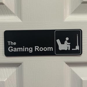 Puede incluir: Un letrero rectangular negro con las palabras "The Gaming Room" en blanco. Un pequeño icono representa a una persona jugando. El letrero está montado en una puerta blanca.