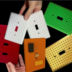 May include: A collection of colorful light switch covers in red, white, green, orange, and gold. The covers have a studded surface and a rectangular opening for the switch. Some have Lego bricks attached.