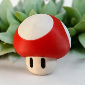 Peut inclure: Une petite figurine de champignon avec un chapeau rouge arrondi surmonté d'un cercle blanc et un pied blanc. Le pied présente deux yeux noirs ovales, simples et verticaux, centrés sur l'avant. L'objet a une finition mate et lisse.