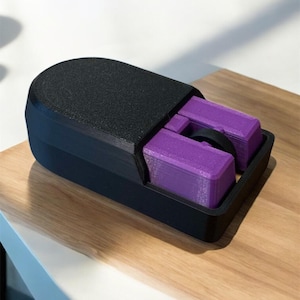 Puede incluir: Un objeto rectangular negro impreso en 3D con una parte superior texturizada y un compartimento que contiene dos bloques rectangulares morados y una pieza circular negra. El objeto est&aacute; sobre una superficie de madera.