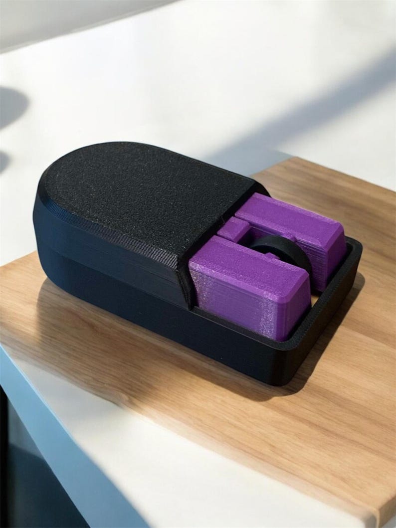 Puede incluir: Un objeto rectangular negro impreso en 3D con una parte superior texturizada y un compartimento que contiene dos bloques rectangulares morados y una pieza circular negra. El objeto est&aacute; sobre una superficie de madera.