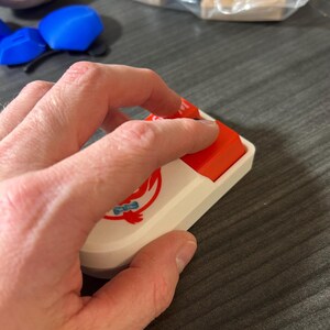 Puede incluir: Una tecla de teclado mec&aacute;nico blanca y roja con un logotipo. La tecla tiene forma rectangular con una base blanca y una parte superior roja. El logotipo es un dise&ntilde;o estilizado en rojo y azul. Una mano est&aacute; presionando la tecla.