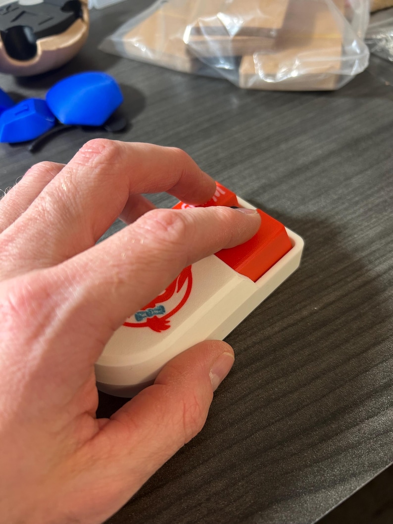 Puede incluir: Una tecla de teclado mec&aacute;nico blanca y roja con un logotipo. La tecla tiene forma rectangular con una base blanca y una parte superior roja. El logotipo es un dise&ntilde;o estilizado en rojo y azul. Una mano est&aacute; presionando la tecla.