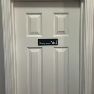 May include: A white, paneled door with a rectangular sign that reads "The Gaming Room." The sign features a graphic of a person seated in a chair with a controller. The door is framed by a white molding.
