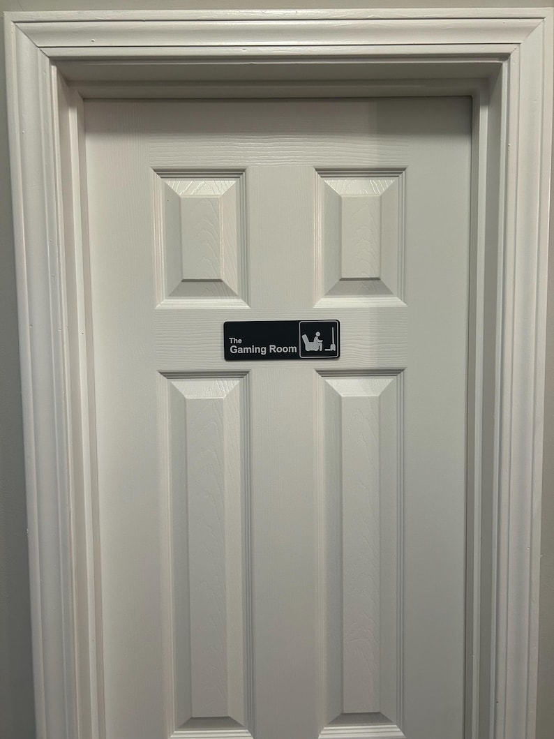 May include: A white, paneled door with a rectangular sign that reads "The Gaming Room." The sign features a graphic of a person seated in a chair with a controller. The door is framed by a white molding.