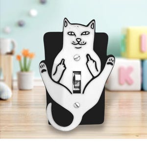 May include: A white and black light switch cover featuring a cat design. The cat is giving the middle finger with both paws. The switch cover is mounted on a black background.