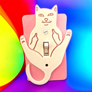 May include: A light pink cat-shaped light switch cover with a white cat design. The cat is giving the middle finger with both hands. The switch is in the "ON" position. The background is a gradient of rainbow colors.