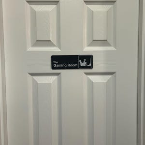 May include: A white door with a black sign that reads "The Gaming Room." The sign features a graphic of a person sitting in a chair playing a video game. The door has a panel design.