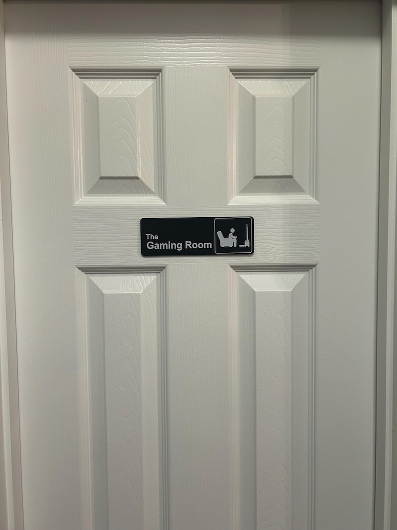 May include: A white door with a black sign that reads "The Gaming Room." The sign features a graphic of a person sitting in a chair playing a video game. The door has a panel design.