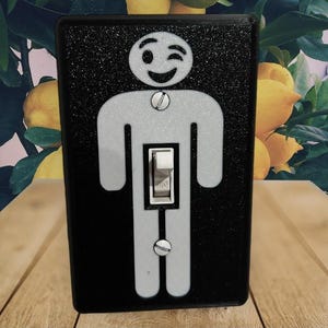 May include: A black light switch cover with a glittery texture, featuring a white outline of a winking stick figure. The switch is in the center of the figure's body, with the word "ON" visible. The background is a wooden surface.