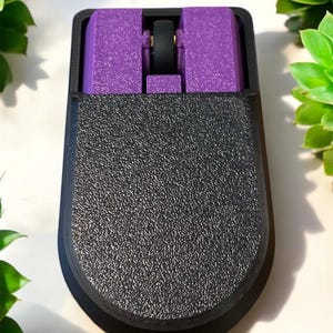 Puede incluir: Un rat&oacute;n de ordenador negro y morado con una superficie texturizada. El rat&oacute;n tiene forma rectangular con la parte inferior redondeada. La secci&oacute;n superior es morada y la rueda de desplazamiento es negra.