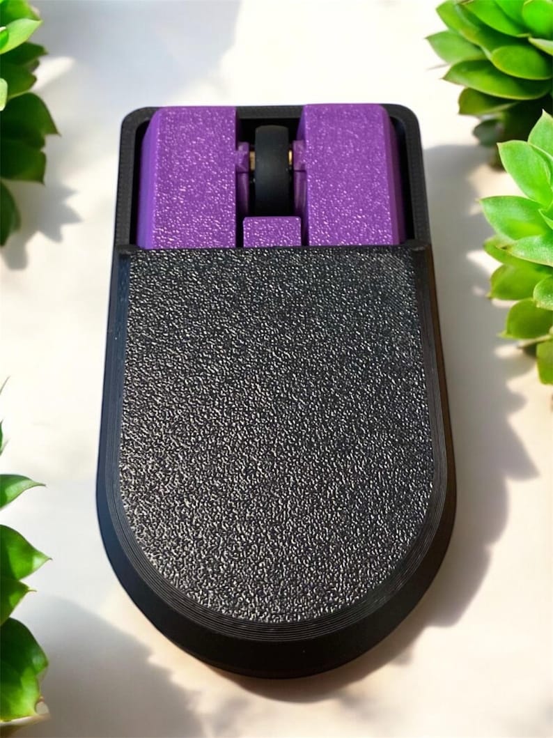 Puede incluir: Un rat&oacute;n de ordenador negro y morado con una superficie texturizada. El rat&oacute;n tiene forma rectangular con la parte inferior redondeada. La secci&oacute;n superior es morada y la rueda de desplazamiento es negra.