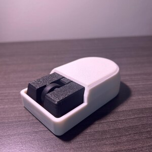 Puede incluir: Un objeto blanco y negro impreso en 3D con una base rectangular y una parte superior curva. El objeto tiene dos bloques rectangulares negros con una peque&ntilde;a rueda negra en el medio. El objeto est&aacute; sobre una superficie de madera.