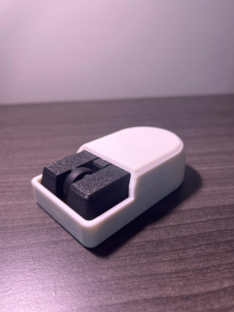 Puede incluir: Un objeto blanco y negro impreso en 3D con una base rectangular y una parte superior curva. El objeto tiene dos bloques rectangulares negros con una peque&ntilde;a rueda negra en el medio. El objeto est&aacute; sobre una superficie de madera.