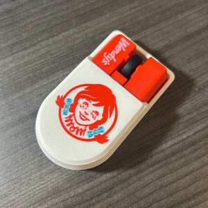 Puede incluir: Un estuche blanco ovalado con el logotipo de Wendy's, que muestra a una ni&ntilde;a pelirroja y la palabra "mom" en azul. El estuche est&aacute; abierto, revelando objetos rojos y negros, con el logotipo de Wendy's visible.