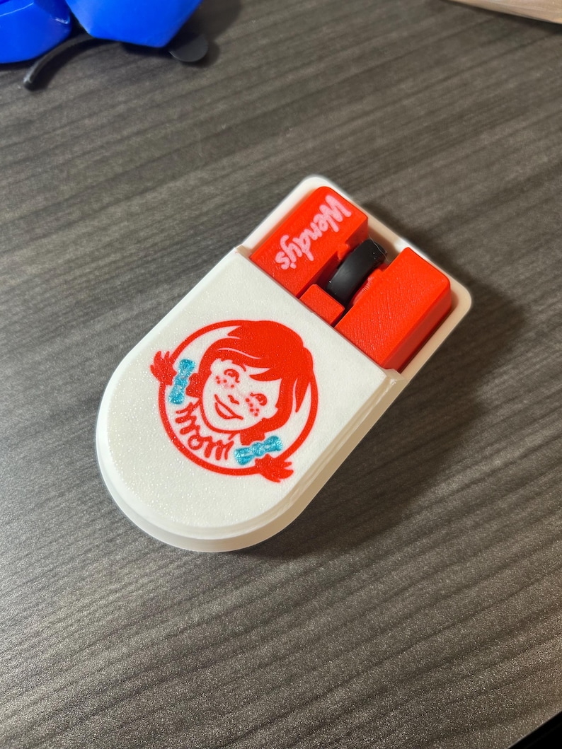 Puede incluir: Un estuche blanco ovalado con el logotipo de Wendy's, que muestra a una ni&ntilde;a pelirroja y la palabra "mom" en azul. El estuche est&aacute; abierto, revelando objetos rojos y negros, con el logotipo de Wendy's visible.