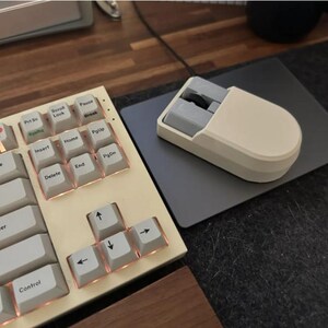 Puede incluir: Un teclado mec&aacute;nico color crema con teclas blancas y bordes color cobre. Un rat&oacute;n color crema a juego se encuentra sobre una alfombrilla gris. El teclado tiene algunas teclas decorativas con im&aacute;genes de una cereza y una flor.