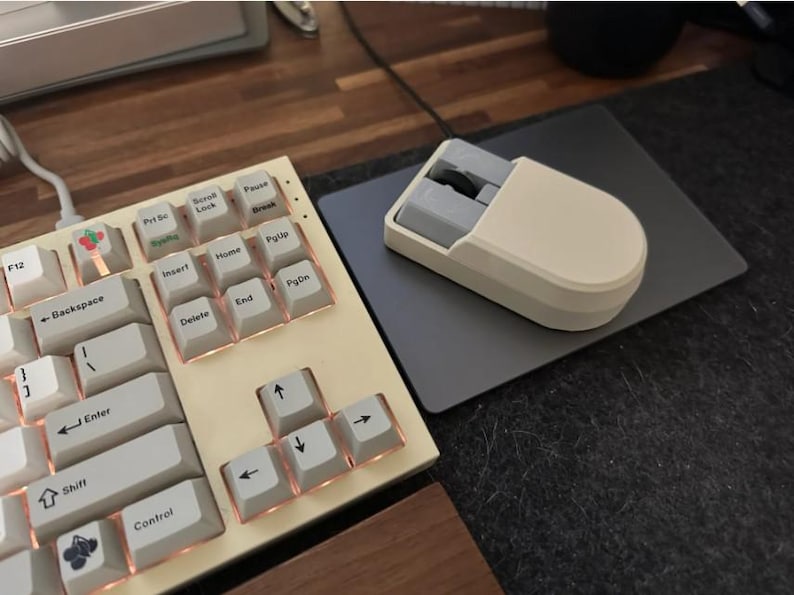Puede incluir: Un teclado mec&aacute;nico color crema con teclas blancas y bordes color cobre. Un rat&oacute;n color crema a juego se encuentra sobre una alfombrilla gris. El teclado tiene algunas teclas decorativas con im&aacute;genes de una cereza y una flor.