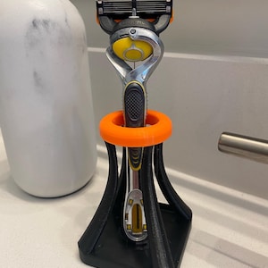 May include: A black and orange razor stand holding a silver, black, and yellow razor. The stand has a square base and a circular top that holds the razor. A white and grey bottle is in the background.