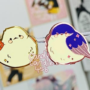 May include: Two enamel pins featuring cartoon birds. One bird has a yellow body with orange and gold accents. The other bird has a yellow body with a blue head and pink accents. The pins have a gold border.