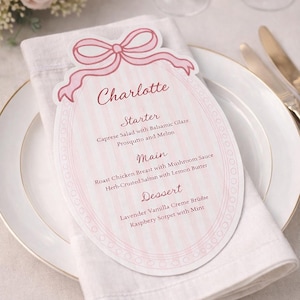Puede incluir: Una tarjeta de menú rosa y blanca con el nombre "Charlotte" en la parte superior, con un diseño de lazo. El menú enumera un entrante, un plato principal y un postre en una servilleta blanca, sobre un plato blanco con borde dorado.