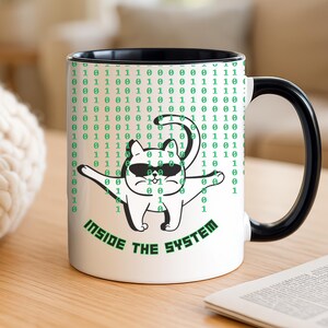 Pode incluir: Caneca de cerâmica branca com alça e borda pretas. A caneca apresenta um gato de desenho animado usando óculos de sol, cercado por código binário verde. O texto na caneca diz "INSIDE THE SYSTEM".