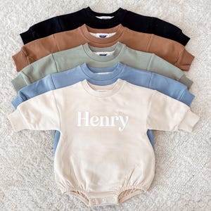 May include: Five baby rompers in a row, each in a different color: black, brown, green, blue, and cream. The cream-colored romper has the name "Henry" embroidered in white. All rompers have long sleeves and a round neckline.