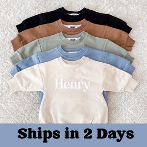 May include: Five crewneck sweatshirts in black, brown, sage green, light blue, and cream. The cream sweatshirt has the name "Henry" embroidered in white. The sweatshirts are arranged in a layered display. The text "Ships in 2 Days" is at the bottom.