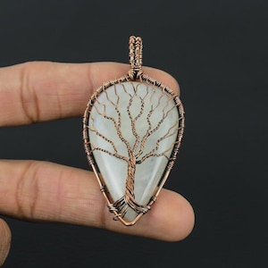 Op de afbeelding: Een druppelvormige hanger met een lichtgekleurde steen en een koperdraad boomontwerp. De takken van de boom strekken zich uit over de steen en de hanger heeft een koperdraad beugel. Een uniek sieraad.