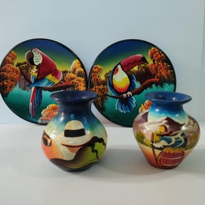 May include: Two round decorative plates featuring colourful parrot and toucan illustrations against a forest background, paired with two small ceramic vases painted with figures wearing hats and carrying walking sticks in a landscape setting.