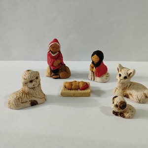 May include: A collection of small ceramic figurines depicting a nativity scene. The figures include a person in a red robe, a person in a black head covering and red robe, a baby in a manger, two sheep, and a small dog. The figurines are beige and brown.