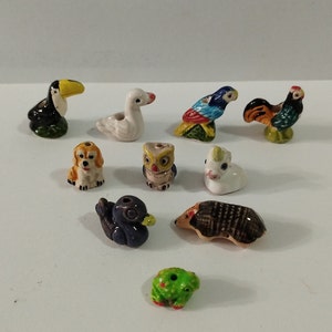 Puede incluir: Una colección de diez pequeñas figuritas de animales de cerámica. Las figuritas incluyen un tucán, un cisne, un loro, un gallo, un perro, un búho, un unicornio, un erizo, una rana y un pájaro. Las figuritas están todas pintadas a mano y tienen colores brillantes.