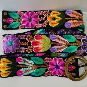 Peut inclure: Une ceinture noire ornée de broderies florales aux couleurs vives, notamment rose, violet, jaune, vert et orange. La ceinture est dotée d'une grande boucle ronde en métal couleur laiton vieilli et de trois boutons-pression en laiton.