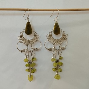 May include: A pair of dangling earrings with a silver-colored filigree design. Each earring features a teardrop-shaped olive-green stone at the top, with a cluster of small, light green stones hanging below. The earrings are suspended from silver-colored hooks.