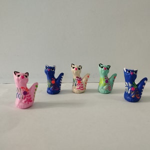 Op de afbeelding: Vijf handgeschilderde keramische kattenbeeldjes. De katten staan in verschillende poses en zijn geschilderd in felle kleuren, waaronder roze, blauw, groen en wit. Elke kat heeft een uniek bloemenmotief.