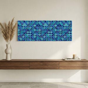 May include: A rectangular wall art piece featuring a pattern of overlapping blue and teal scales. The artwork hangs above a dark wood floating shelf. A white vase with dried plants and a candle are also visible.