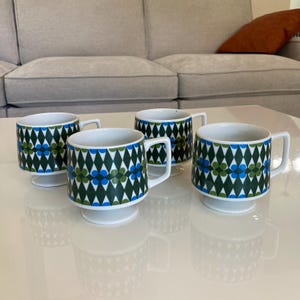 Può includere: Quattro tazze vintage in ceramica con un motivo geometrico verde, blu e bianco. Le tazze hanno una base e un manico bianchi. Il motivo presenta forme a diamante e accenti floreali. Le tazze sono disposte su una superficie bianca.