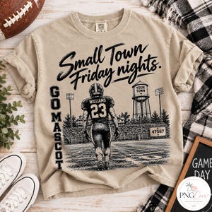 Puede incluir: Una camiseta beige de manga corta con un gráfico negro de un jugador de fútbol americano con el número 23 en un campo. El diseño incluye una torre de agua, luces de estadio y el texto Small Town Friday nights y GO MASCOT.