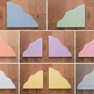 Op de afbeelding: Een collectie van acht paar houten wandplanken, elk met een opvallende, golvende, wolkachtige bovenrand. De planken zijn afgewerkt in verschillende matte kleuren, waaronder blauw, groen, oranje, paars, roze, wit, geel en natuurlijk hout.