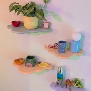 Peut inclure: Présentation de plusieurs étagères en forme de nuage, chacune ornée d'objets décoratifs originaux. On y trouve une plante en pot, des petits bols, une figurine en forme de pain grillé et d'autres objets insolites. La palette de couleurs est composée de pastels et d'accents vifs.