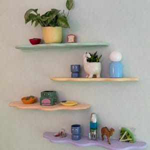 Op de afbeelding: Decoratieve wolkvormige planken in pastelkleuren, met diverse items. De planken tonen een gele potplant, een rode kom, een toastfiguur, een blauw beeldje, een lama-planter en een raket. Afmetingen in cm.