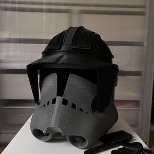 Könnte beinhalten: Ein grau-schwarzer Helm mit Visier und abgerundeter Oberseite. Der Helm hat ein detailliertes Design mit verschiedenen Komponenten, darunter ein Visier und zusätzliche schwarze Teile. Der Helm ist wahrscheinlich ein Kostüm oder eine Requisite.