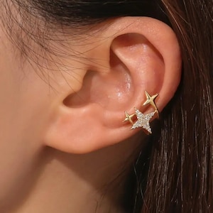 Könnte beinhalten: Goldfarbene Ohrklemme mit drei Sterndesigns. Ein Stern ist mit kleinen, klaren Kristallen besetzt, die anderen beiden sind massiv. Der Schmuck wird am Ohr getragen.