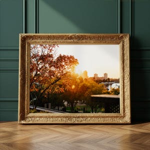 May include: A framed landscape photograph featuring a city skyline at sunset. The ornate gold frame surrounds a view of trees with autumn foliage, a bridge, and buildings bathed in warm sunlight. The frame rests on a wooden floor.