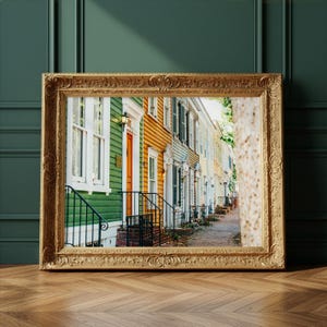 May include: A framed print of colorful historic houses. The houses are painted in shades of green, orange, and yellow, with white trim and black railings. The print is displayed in a gold ornate frame, resting on a wooden floor.