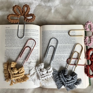 May include: A collection of handmade macrame bookmarks in various colors, including tan, white, gray, pink, and red. Each bookmark features a unique design, such as a flower or a tassel, attached to a metal paperclip. The bookmarks are displayed on an open book.